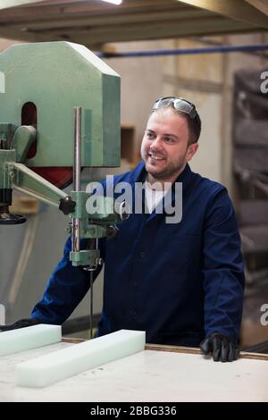 Process of furniture production. A young man in a dark coat cut the foam for the sofa on a cutting machine. He is wearing a safety glasses and gloves. Stock Photo