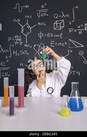 child scientist with glasses in lab coat looking a flask with chemical liquid, on blackboard background with science formulas, concept of back to scho Stock Photo