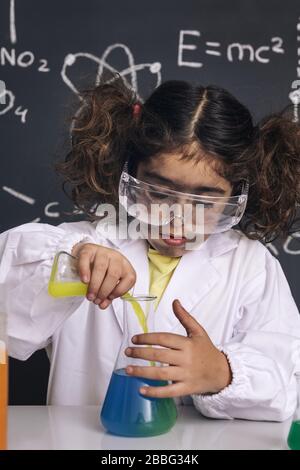 scientist kid with goggles and gloves in lab coat mixing chemical liquids in flasks, blackboard background with science formulas, explosion in the lab Stock Photo
