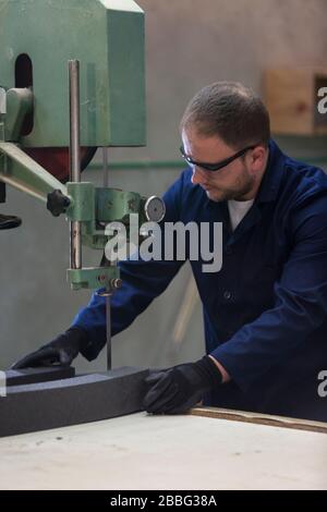 Process of furniture production. A young man in a dark coat cut the foam for the sofa on a cutting machine. He is wearing a safety glasses and gloves. Stock Photo