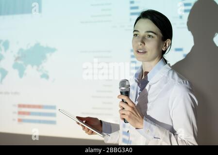 Young woman in lab coat using thesis plan on tablet while presenting project at medical convention Stock Photo