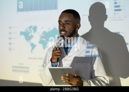 Confident young Black doctor in lab coat using tablet and microphone while giving speech at scientific conference Stock Photo
