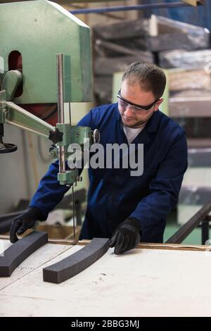 Process of furniture production. A young man in a dark coat cut the foam for the sofa on a cutting machine. He is wearing a safety glasses and gloves. Stock Photo
