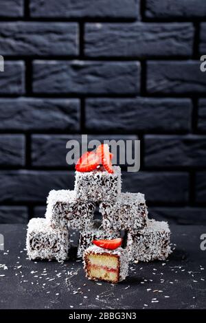 Lamingtons vanilla sponge mini cakes with strawberry jam layer inside, coated with chocolate and coconut on top on a dark concrete table in front of a Stock Photo