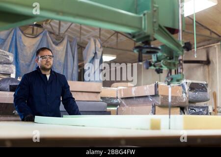 Process of furniture production. A young man in a dark coat cut the foam for the sofa on a cutting machine. Behind him there are a lot of finished sof Stock Photo