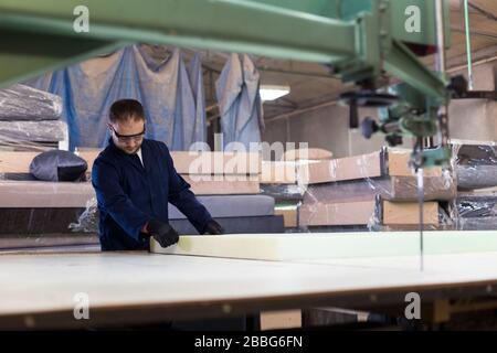 Process of furniture production. A young man in a dark coat cut the foam for the sofa on a cutting machine. Behind him there are a lot of finished sof Stock Photo