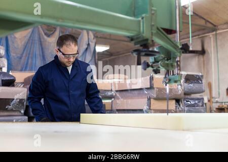 Process of furniture production. A young man in a dark coat cut the foam for the sofa on a cutting machine. Behind him there are a lot of finished sof Stock Photo