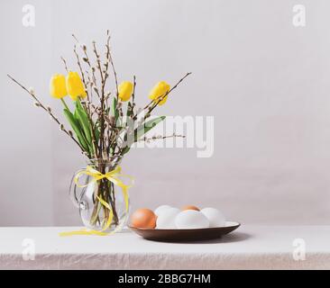 Festive Easter eggs in a clay plate a vase with yellow tulips and willow branches on the table. Happy Easter concept. Stock Photo
