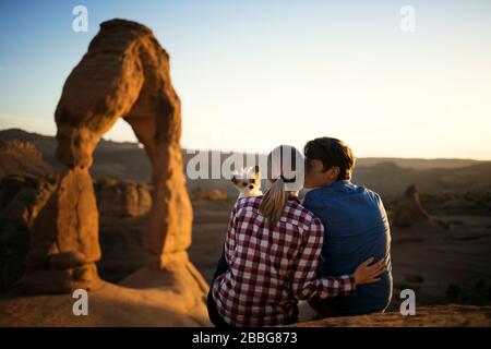 Young man kissing his girlfriend on the cheek while their dog looks over her shoulder with a scenic canyon in the background Stock Photo