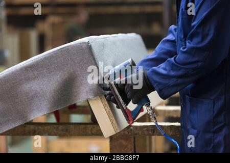 Process of furniture production. A young man in a dark coat who puts together one part of the sofa with a stapler on a working desk. He has a black sa Stock Photo