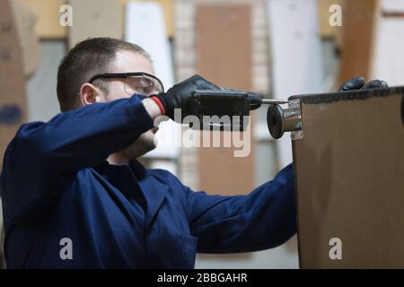 Process of furniture production. A young man in a dark coat attaches the sofa leg with a cordless drill. Behind him on are a lot of tools and material Stock Photo