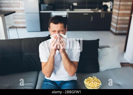 Young man watch tv in his own apartment. Guy sneezing into white napkin. Sick and ill at home. Suffer from headache or nose pain. Serious disease and Stock Photo