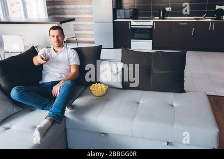 Young man watch tv in his own apartment. Joyful happy attractive ordinary guy hold remote control with straightened hand. Switching tv channels Stock Photo