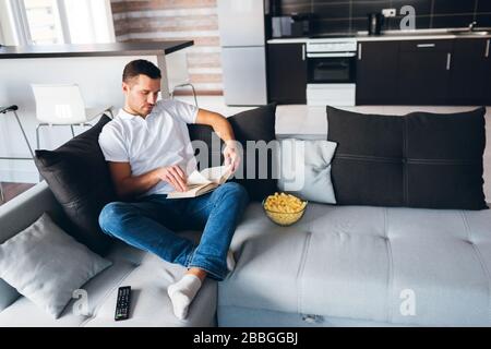 Young man watch tv in his own apartment. Calm peaceful concentrated guy sit on couhc and read book after watching movie. Reading in silence. Studying Stock Photo
