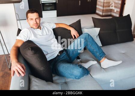 Young man watch tv in his own apartment. Nice cheerful confident guy posing on camera and smile. Sit on comfortable couch and posing. Alone in room Stock Photo