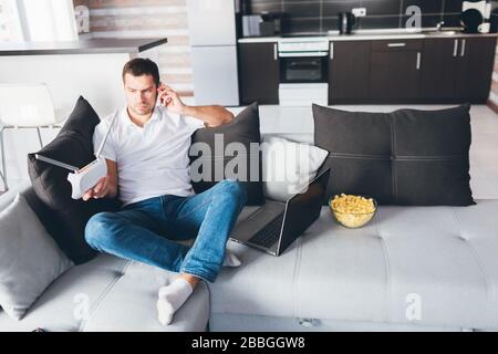 Young man watch tv in his own apartment. Guy sit on couch and talking on phone. Hold wi-fi router in hand and look at it. Customising internet device Stock Photo