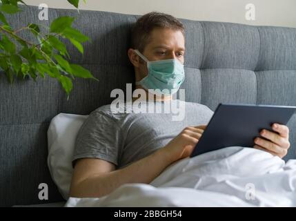Sick man with medical mask using tablet pc in bed. Home quarantine concept. Stock Photo