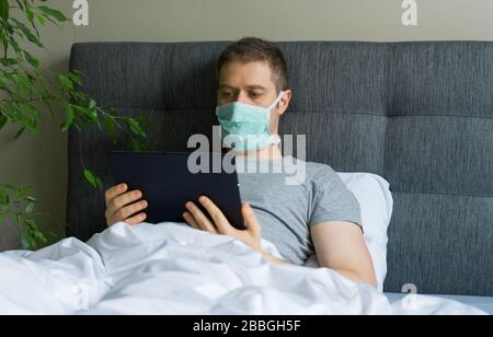 Sick man with medical mask using tablet pc in bed. Home quarantine concept. Stock Photo