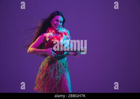 Fabulous Cinco de Mayo female dancer on purple studio background in neon light. Beautiful female model with ukulele, Mexico, Hawaii style. Celebration, holiday, music, beauty and fashion concept. Stock Photo