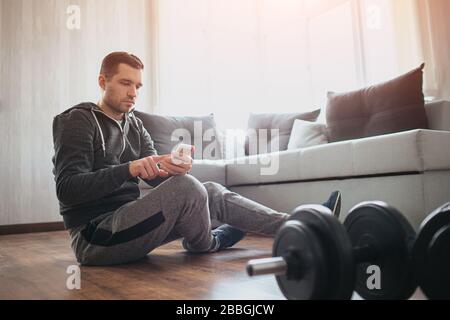 Young ordinary man go in for sport at home. Has rest with sitting on floor and use smartphone. Freshman in workout relaxing after training. Alone in Stock Photo