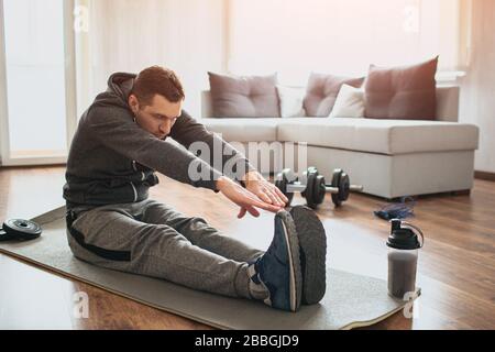 Young ordinary man go in for sport at home. Beginner or amateur in workout activity sit on mat and stretch to his toes. Hardworking guy exercising in Stock Photo