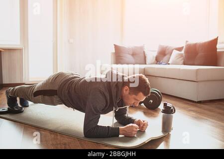 Young ordinary man go in for sport at home. Full size pictrue of freshman in workout and beginner guy stand in plank position on mat. Look down and Stock Photo