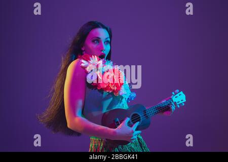 Fabulous Cinco de Mayo female dancer on purple studio background in neon light. Beautiful female model with ukulele, Mexico, Hawaii style. Celebration, holiday, music, beauty and fashion concept. Stock Photo