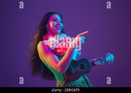 Fabulous Cinco de Mayo female dancer on purple studio background in neon light. Beautiful female model with ukulele, Mexico, Hawaii style. Celebration, holiday, music, beauty and fashion concept. Stock Photo