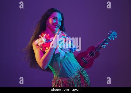 Fabulous Cinco de Mayo female dancer on purple studio background in neon light. Beautiful female model with ukulele, Mexico, Hawaii style. Celebration, holiday, music, beauty and fashion concept. Stock Photo