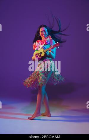 Fabulous Cinco de Mayo female dancer on purple studio background in neon light. Beautiful female model with ukulele, Mexico, Hawaii style. Celebration, holiday, music, beauty and fashion concept. Stock Photo