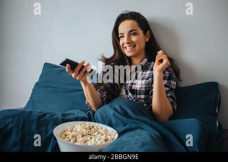 Young beautiful woman in morning bed at home. Positive nice cheerful person watching tv or movie. Enjoying time in bed and eating popcorn. Stock Photo
