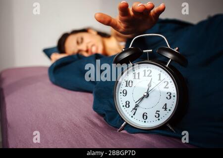 Young beautiful woman in morning bed at home. Irritated by clock ringing. Reaching it with hand. Time to wake up. Stock Photo