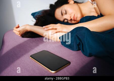 Young beautiful woman in morning bed at home. Sleeping and resting. Sun shines inside. Smartphone lying on ned besides woman. Stock Photo