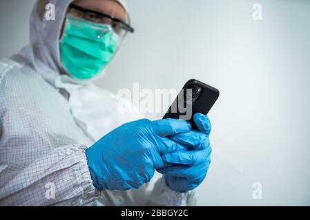 Caucasian man wearing a protective suit and face mask using smartphone on laboratory. Doctor using phone device. Coronavirus has caused emergency situ Stock Photo
