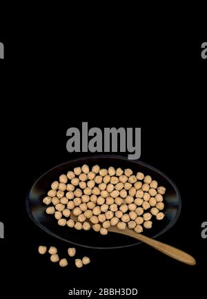 Dried Kabuli chickpeas, also called Garbanzo beans presented in a black oval bowl with a brown bamboo spoon, on a dark background. Stock Photo