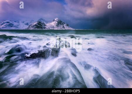 Big waves washing over rocks on the beach with dark storm clouds and snowcapped mountains on the horizon Stock Photo