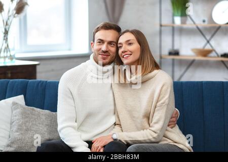 Wife and husband sitting at sofa, smiling, looking at camera. Positive married couple. Spouses spend free time together at home. Happy young newlyweds Stock Photo