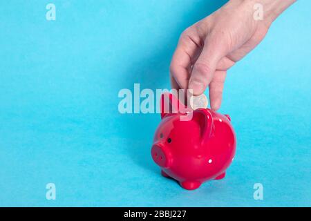 Male hand throws one euro into a red piggy bank in a white dot on a blue background. Cocept. Copy space. Stock Photo