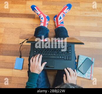 Man wearing union jack flag sock sitting on wooden floor with laptop. Working from home, self isolation, social distancing, Coronavirus... concept Stock Photo