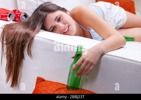 Young intoxicated teenage girl lying on a sofa holding an empty green beer bottle over the side with crumpled cans beside her in a concept of alcohol Stock Photo