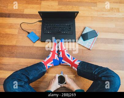 Man wearing union jack flag sock sitting on wooden floor with laptop. Working from home, self isolation, social distancing, Coronavirus... concept Stock Photo