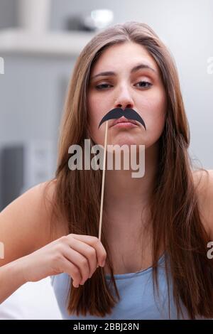 Young teenage girl goofing around with party accessories holding a mustache to her upper lip with a supercilious expression, in close up indoors Stock Photo
