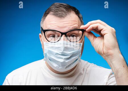Close up portrait of senior male wearing safety mask posing in glasses isolated at blue Stock Photo