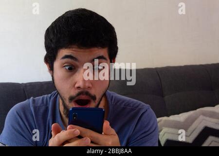 Bearded Hispanic young man in a living room using his smartphone and showing negative reactions such as anger, frustration, boredom, anger and other c Stock Photo