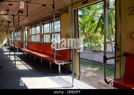 Interior of empty suburban train on the move. Tropical palm trees through open doors. Orange hard seats with no people and metal handrails. Sri Lanka Stock Photo