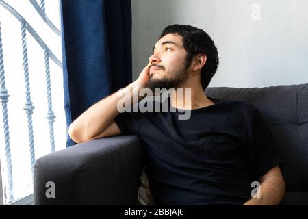 Bearded Hispanic young man sitting on a sofa very thoughtful looking out a window Stock Photo