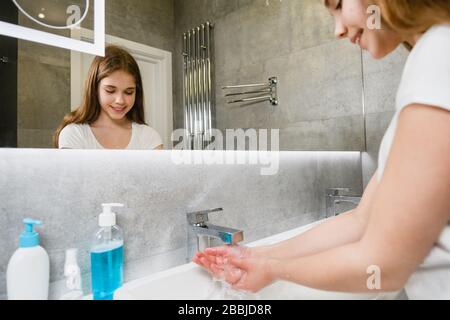 Child in bathroom washing hands and arms with liquid soap. Virus protection concept. A smiling girl is reflected in the bathroom mirror while washing Stock Photo