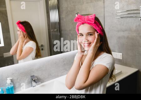 Young smiling woman in pink hair headband bow remove makeup and wash skin in bathroom, hygiene home ritual, lather hands and cheeks Stock Photo