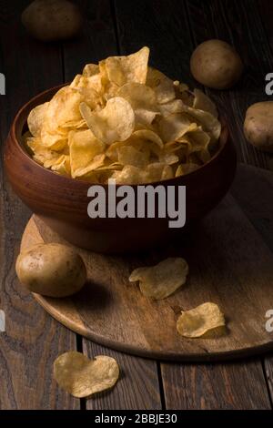 Potato chips in a bowl on a rustic wooden background with whole potatoes. Salty crisps scattered on a table. Stock Photo