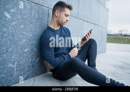 Side view of modern male in casual wear sitting near stone wall and browsing smartphone while resting on city street Stock Photo